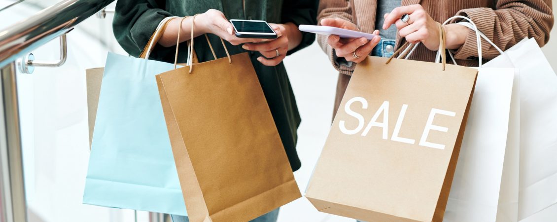 hands-of-two-young-contemporary-shoppers-with-smartphones-and-paperbags-scrolling-through-online-goods-and-comparing-prices